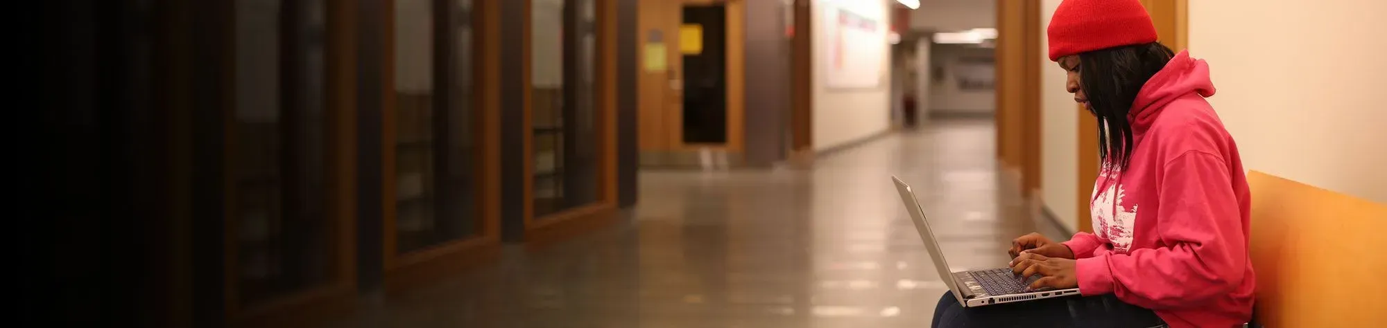 A young woman in a red hoodie sitting in a school hall while using her laptop.